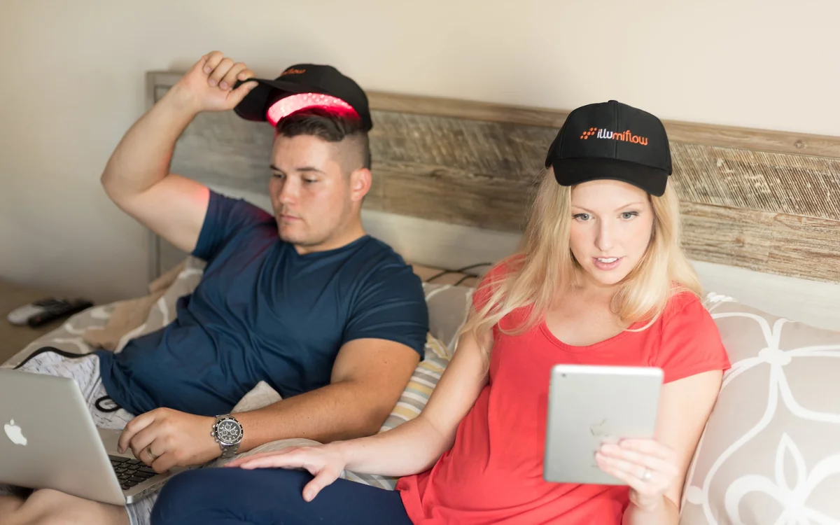 Two people wearing Illumiflow laser caps at home, one cap lifted to show the red lasers